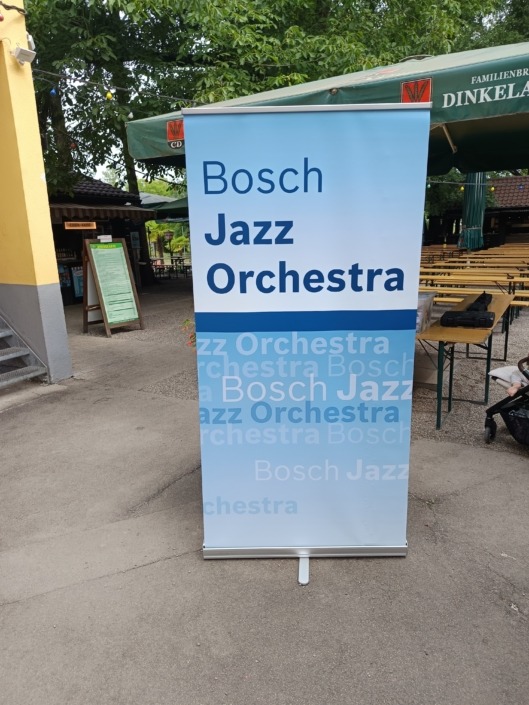 Ein Roll-Up mit der Aufschrift Bosch Jazz Orchestra steht auf einem Gehweg neben Bierbänken und Bäumen.