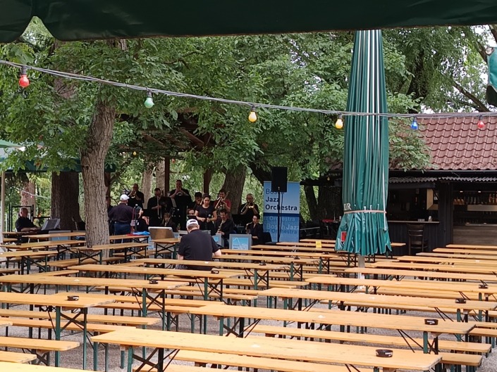 Eine Jazzband spielt in einem Biergarten unter Bäumen, umgeben von leeren Holzbänken und bunten Lichterketten.