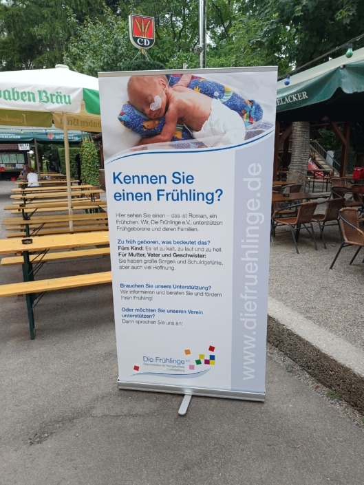 Ein Roll-Up mit einem Foto eines Frühgeborenen und Text zur Unterstützung von Frühgeborenen steht auf einem Biergartenweg.