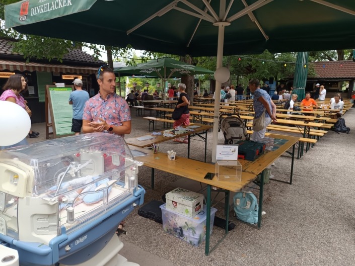 Ein Mann steht neben einem Inkubator unter einem grünen Sonnenschirm in einem Biergarten mit leeren Bänken.