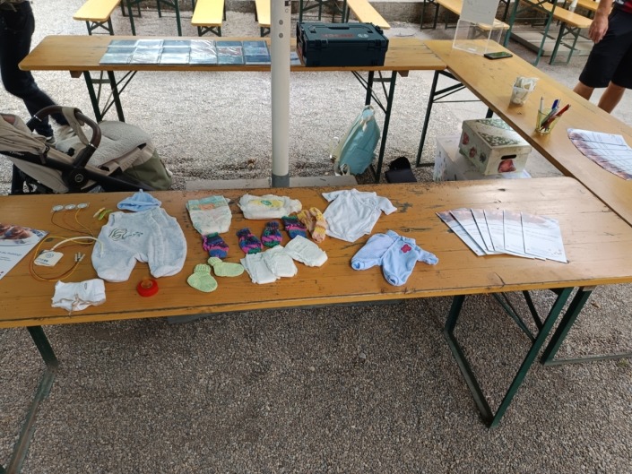 Babybekleidung und Infomaterialien liegen auf Holztischen im Freien.
