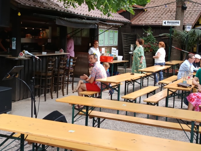 Ein Biergarten mit Holzbänken, Menschen sitzen und stehen, ein Kiosk im Hintergrund, Schild Ausgang zum Neckar.