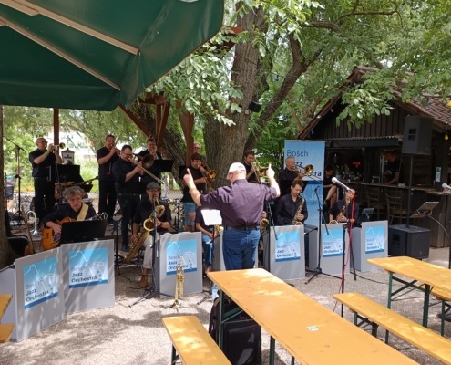 Eine Jazzband spielt unter Bäumen, dirigiert von einem Mann, mit leeren Bänken im Vordergrund.