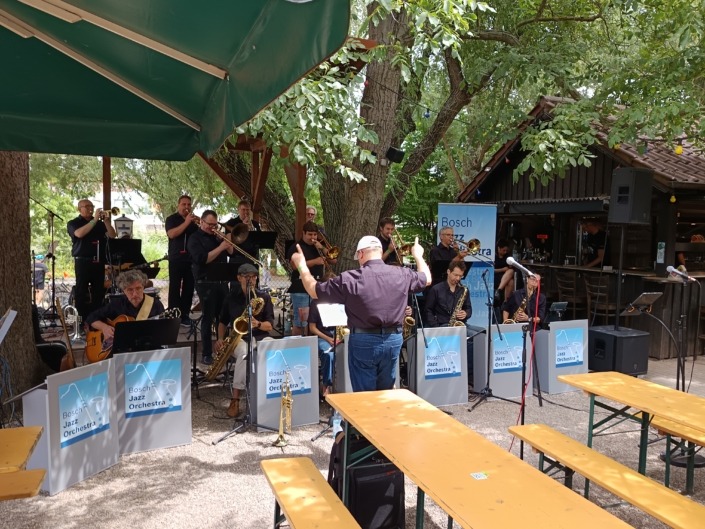 Eine Jazzband spielt unter Bäumen, dirigiert von einem Mann, mit leeren Bänken im Vordergrund.