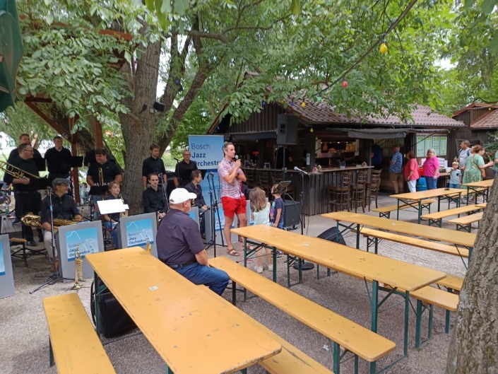 Eine Jazzband spielt im Freien neben Bierbänken, umgeben von Bäumen und Zuschauern.