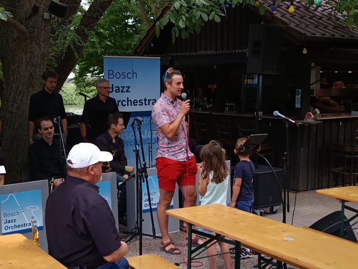 Ein Mann spricht in ein Mikrofon, umgeben von Musikern und Kindern, vor einem Banner des Bosch Jazz Orchestra.