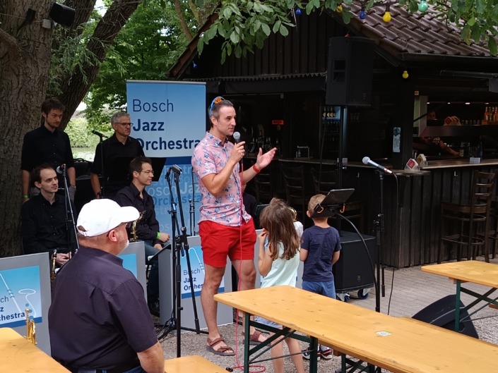 Ein Mann spricht vor einer Jazzband im Freien, während Kinder zuhören. Im Hintergrund eine Bar und Bäume.