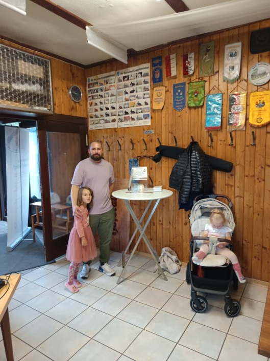 Mann und Mädchen stehen neben einem Tisch; Baby im Kinderwagen, Holzwand mit Postern im Hintergrund.