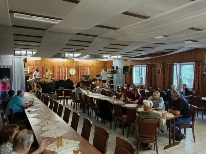 Menschen sitzen in einem großen Raum mit Holzwänden an langen Tischen, während Musiker auf einer Bühne spielen.