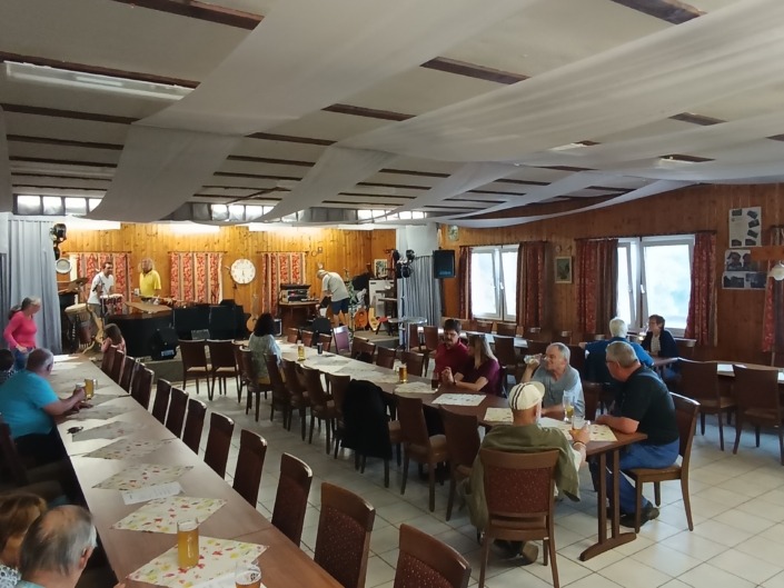 Menschen sitzen in einem Saal mit Holzwänden, während eine Band auf der Bühne probt. Tische sind mit Getränken gedeckt.