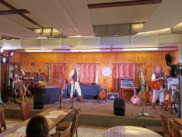 Eine Band probt auf einer Holz-Bühne mit Instrumenten, darunter Gitarren und Schlagzeug, vor einem Publikum in einem Saal.