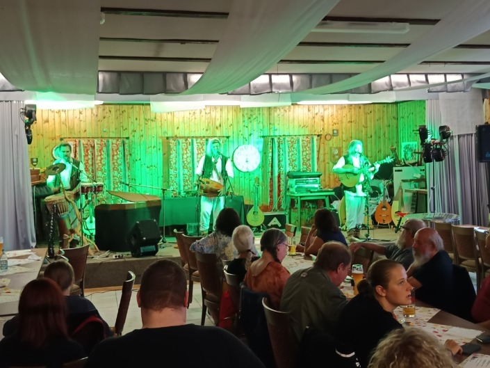 Drei Musiker spielen auf einer Bühne vor einem sitzenden Publikum in einem beleuchteten Raum mit Holzwänden.