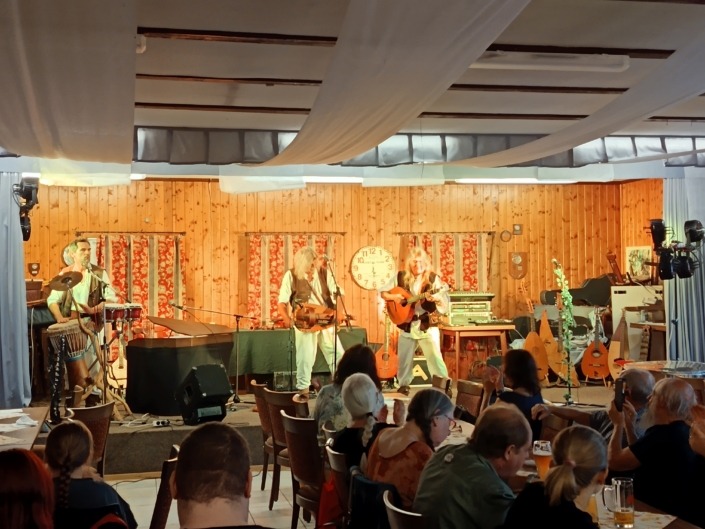 Eine Band spielt auf einer Holzbühne, umgeben von Instrumenten, vor einem Publikum in einem dekorierten Saal.