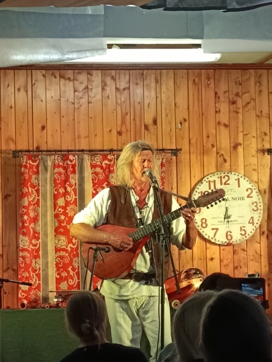 Ein Musiker spielt Gitarre auf einer Bühne mit Holzverkleidung und rotem Vorhang. Eine große Uhr hängt an der Wand.