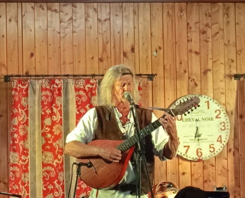 Ein Musiker mit Gitarre singt auf einer Holzbühne, im Hintergrund eine Uhr und rote Vorhänge.
