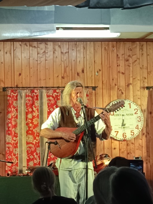 Ein Musiker mit Gitarre singt auf einer Holzbühne, im Hintergrund eine Uhr und rote Vorhänge.