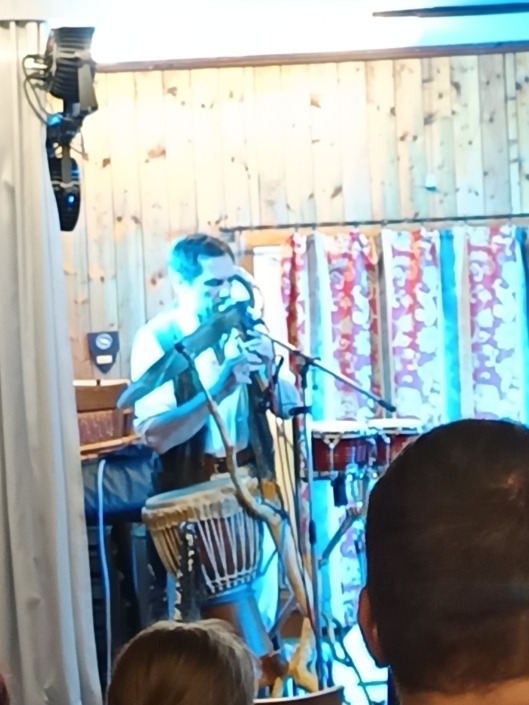 Ein Musiker spielt Flöte und Trommel auf einer Bühne mit Holzwand und bunten Vorhängen.