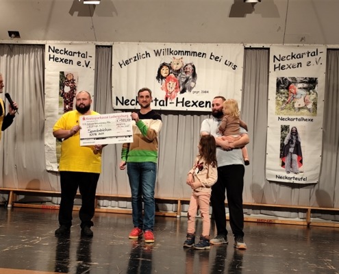 Eine Gruppe von Menschen auf einer Bühne hält einen großen Scheck vor Bannern mit der Aufschrift Neckartal-Hexen.
