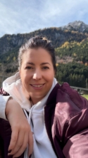 Eine Person in Outdoor-Kleidung lächelt vor einer Berglandschaft mit herbstlichen Bäumen.